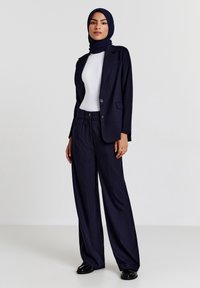 Marineblaues Nadelstreifenanzug mit einem taillierten Blazer und weit geschnittenen Hosenbeinen, kombiniert mit einem weißen Oberteil und passendem Hijab. Glatte Textur und maßgeschneiderte Passform.