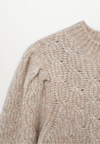Pull en maille beige épaisse avec col montant côtelé et motifs torsadés sur les épaules et le corps.