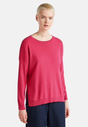 Felle roze gebreide trui met een ronde hals, lange mouwen en zijsplitten, gecombineerd met donkerblauwe broeken. De stof heeft een gladde textuur.