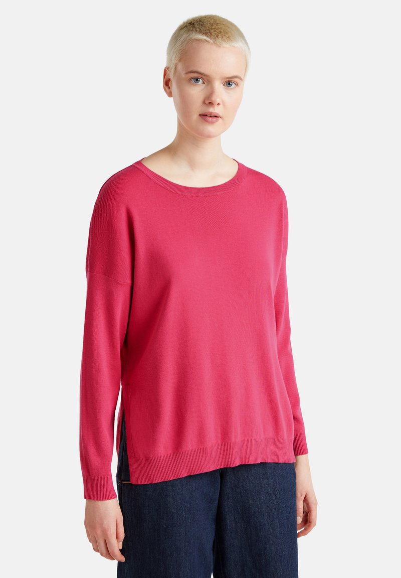 Maglione lavorato a maglia fucsia con scollatura rotonda, maniche lunghe e spacchi laterali, abbinato a pantaloni blu scuro. Il tessuto ha una texture liscia.