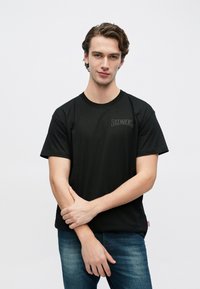 Must lühikeste varrukatega T-särk ümmarguse kaelusega, millel on ülemises vasakus nurgas peen "DIESEL" logo ning mis on kantud siniste teksadega.