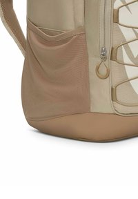 Mochila beige que presenta un bolsillo de malla texturizada, cierre de cremallera y un detalle metálico circular. La sección inferior tiene un acabado liso y sólido en tono marrón claro.