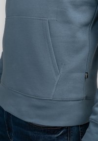 Modrá mikina z bavlněné směsové látky, s klokaní kapsou, žebrovaným lemem a texturovanými švy. Ozdobená detailním prošitím.