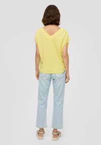 Gelbes Kurzarmoberteil mit V-Ausschnitt, kombiniert mit hellblauen geraden Jeans. Lässiger Stoff mit insgesamt lockerer Passform.