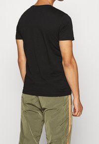 Mann trägt ein schlichtes schwarzes T-Shirt mit kurzen Ärmeln und olivgrüne Hosen mit orangefarbenen Seitenstreifen, steht mit dem Rücken zur Kamera.