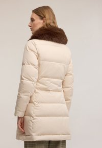 Pikowany płaszcz w jasno beżowym kolorze z brązowym sztucznym kołnierzem z futra. Posiada poziome przeszycia, długie rękawy oraz długość sięgającą poniżej bioder.