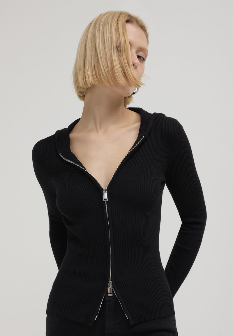 Schwarze, gerippte Kapuzenjacke mit Reißverschluss, V-Ausschnitt, langen Ärmeln und silbernen Reißverschlussdetails. Sie hat ein figurbetontes Design und einen strukturierten Stoff.