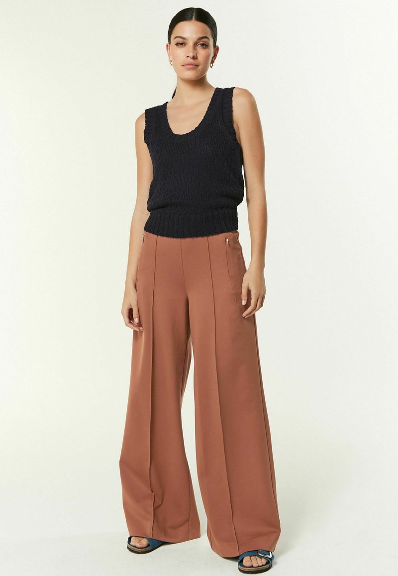 Débardeur sans manches tricoté bleu marine associé à un pantalon large taille haute orange-brun. Les sandales ont une lanière bleue et une semelle plate.