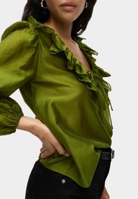 Groene, lichte blouse met een ruches halslijn en mouwen, met een strikdetail aan de voorkant. Gecombineerd met zwarte broek en een riem.
