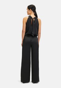 Svart ärmlös jumpsuit med en knytning bak, vid byxben och en mjuk tygkvalitet, som framhäver en minimalistisk silhuett.