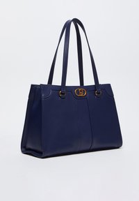 Borsa tote in pelle blu navy con due manici lunghi, hardware dorato e un design strutturato e liscio. Presenta una chiusura decorativa centrale.