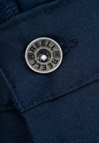 Navy fabric with a textured weave, featuring a silver metal button engraved with "REELL" and a central stud. Close-up detail on the button.