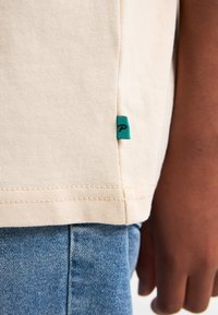Beige shirtzoom met een klein groen label bovenop blauwe denim jeans en een hand van een persoon rustend naast de heup.