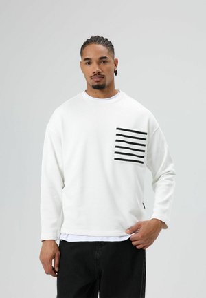 Witte trui met lange mouwen en een grote voorzak met zwarte horizontale strepen. Gecombineerd met zwarte jeans.