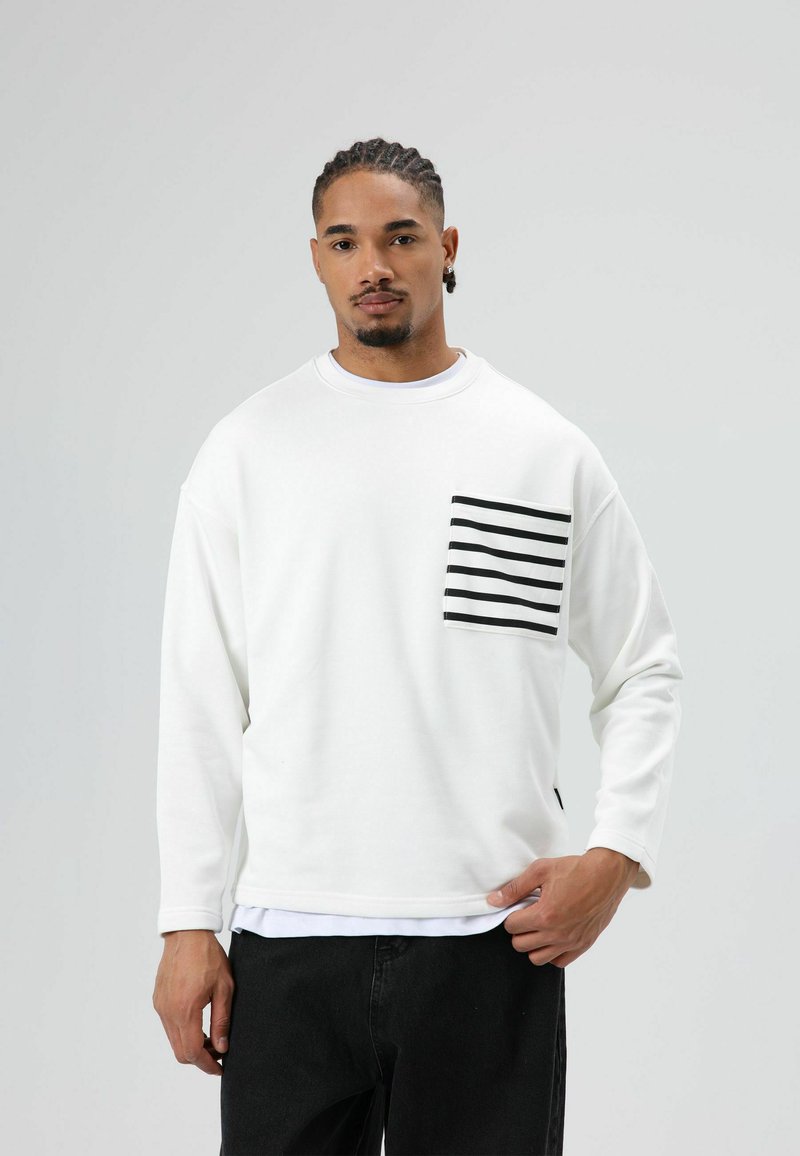 Witte trui met lange mouwen en een grote voorzak met zwarte horizontale strepen. Gecombineerd met zwarte jeans.