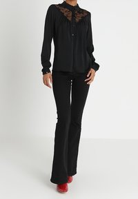 Femme portant un chemisier noir à manches longues avec des détails en dentelle, un pantalon noir évasé et des bottes rouges à talons, posant devant un fond blanc uni.