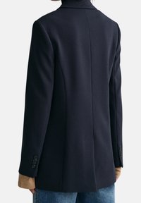 Blazer in misto lana navy con colletto strutturato, chiusura con un solo bottone e dettagli a manica a fantasia. Presenta una vestibilità sartoriale e spalline.