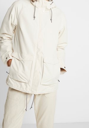 Persoon gekleed in een gebroken witte capuchonjas met grote voorkantzakken en trekkoordjes, gecombineerd met een bijpassende gebroken witte broek. Handen in de zakken.