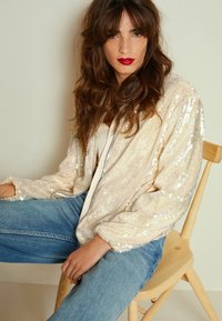 Paljetbombjakke i lys beige med en lang, løs pasform; har en sølvfarvet lynlås og elastiske manchetter, parret med blå denim jeans.