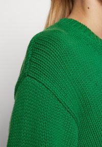 Gros plan sur l'épaule et le haut du bras d'un pull vert en laine porté par une personne à la peau claire et aux cheveux blonds.