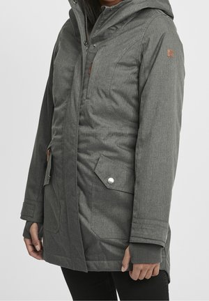 Femme portant une longue veste d'hiver grise à capuche avec fermeture éclair à l'avant et poches à boutons-pression, debout les mains détendues le long du corps.