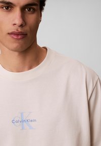 Νεαρός άντρας που φοράει μπέζ μπλουζάκι Calvin Klein με μπλε λογότυπο στο στήθος, σε απλό ανοιχτό γκρι φόντο.