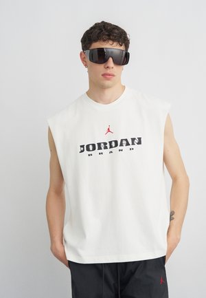 Junger Mann mit übergroßen schwarzen Sonnenbrillen, weißem ärmellosem Jordan Brand Shirt und schwarzen Hosen, steht vor einfachem grauen Hintergrund.