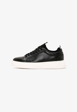 Zapatilla negra de cuero con puntera redondeada, diseño de cordones y suela de goma blanca. Textura suave con detalles mínimos y lengüeta en el talón.