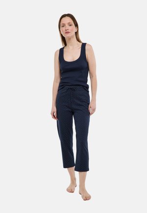 Femme debout pieds nus portant un débardeur bleu marine et un pantalon court bleu marine à pois blancs, sur un fond uni.