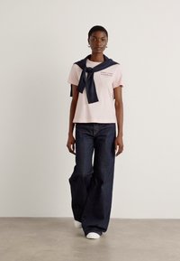 T-shirt rose clair avec logo, porté sur un pull bleu marine noué au cou ; associé à un jean large bleu foncé et des baskets blanches.