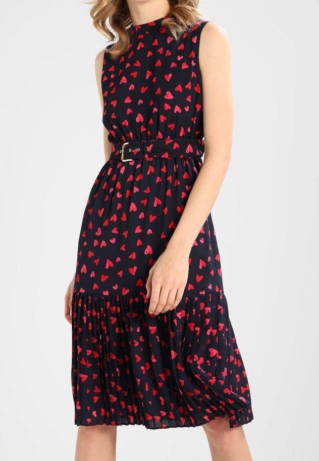 Robe midi noire sans manches avec motif de cœurs rouges, taille ceinturée, jupe plissée, portée par une personne debout devant un fond uni.