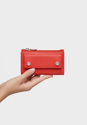 Main tenant un portefeuille en cuir rouge avec une poche avant à pression et une fermeture éclair supérieure sur un fond blanc uni.