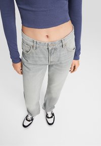 Jeans gris claro con un diseño de pierna recta y cinco bolsillos; el modelo lleva una camiseta de manga larga azul marino y zapatillas en blanco y negro.