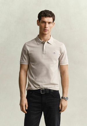 Jeune homme portant un polo beige clair, un pantalon noir avec une ceinture et une montre-bracelet argentée, debout devant un fond clair uni.