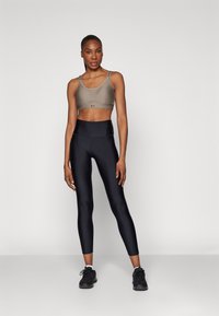 Idrettsleder kvinne står, iført grå sports-BH, svarte høye leggings, svarte joggesko, mot en enkel lys bakgrunn.