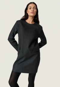 Kátrány csíkos ruha hosszú ujjakkal, kerek dekoltázzsal és oldalsó zsebekkel; textúrált anyagból készült, enyhén testhezálló fazon.