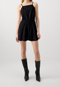 Mono de lino negro con tirantes finos y cinturilla elástica, combinado con botas negras hasta la rodilla con textura acanalada y punta afilada.