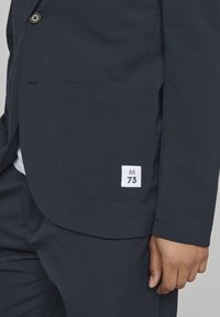 Tamsiai mėlynos spalvos švarkas ir kelnes, dėvimos žmogaus, švarko priekinėje kišenėje matoma balta etiketė, pažymėta "M 73".