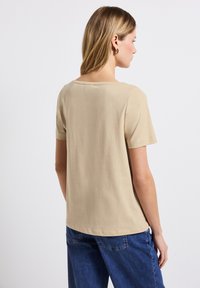 Beige tričko s krátkým rukávem a kulatým výstřihem, hladkou texturou a uvolněným střihem, v kombinaci s modrými džínami.