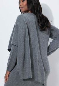 Suéter de punto gris claro con un diseño relajado, que presenta un diseño drapeado y mangas largas. Textura suave con acentos mínimos.