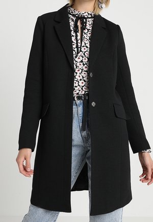 Vrouw draagt een zwarte jas met knopen over een bloemenblouse met striksluiting, gecombineerd met lichtblauwe jeans en grote creolen.