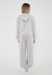 Felpa oversized grigia chiara con cappuccio regolabile, abbinata a pantaloni della tuta a gamba larga coordinati. Realizzata in tessuto morbido, con polsini elasticizzati.