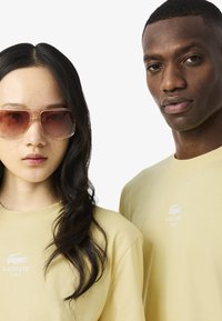 Due persone che indossano t-shirt di Lacoste Paris di un giallo pallido, una con lunghi capelli neri e occhiali da sole colorati, rivolte in avanti contro uno sfondo bianco.