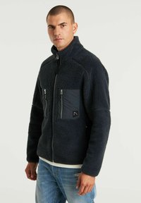 Giacca in pile blu navy con colletto alto, tasche sul petto con zip e tessuto testurizzato. Presenta un pannello frontale a contrasto e una vestibilità affusolata.