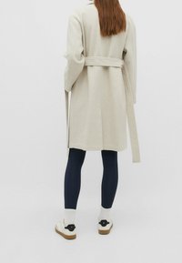 Beżowy wełniany płaszcz z paskiem do wiązania, długimi rękawami i prostym krojem. W zestawie granatowe legginsy i białe sneakersy z czarnymi akcentami.