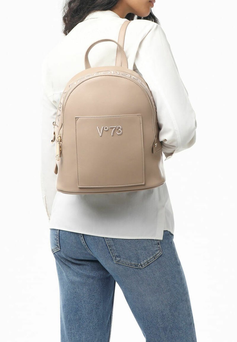 Zaino in finta pelle beige con forma arrotondata, dettagli in metallo dorato, logo frontale e spallacci regolabili, indossato sulla schiena.