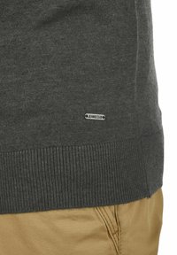 Pull gris foncé en tricot, avec un ourlet côtelé et une petite étiquette de logo. Associé à un pantalon beige clair.