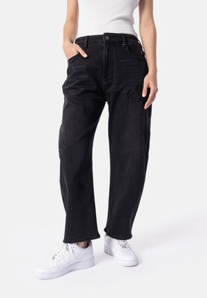 Persona che indossa jeans neri larghi con sottili toppe testurizzate, top bianco senza maniche e sneaker bianche, in piedi contro uno sfondo semplice.