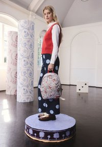 Szőke nő piros mellényben és fehér ingben, virágmintás hátizsákot tartva, festett kör alakú platformon áll egy világos szobában, mintás oszlopokkal.