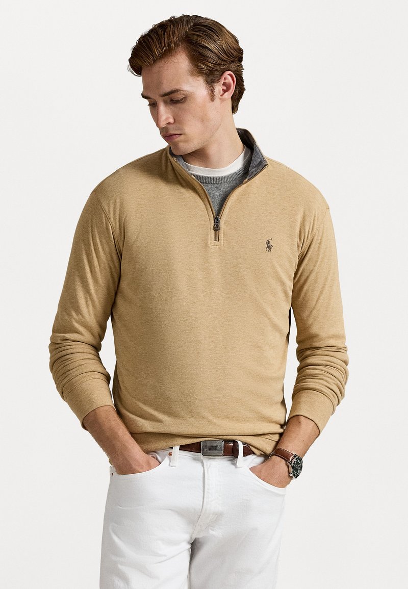 Šviesiai smėlio spalvos zip-up megztinis su pilku vidiniu apykakle, ilgomis rankovėmis, grublėtomis rankogaliais ir logotipu ant krūtinės, vilkimas virš baltos marškinėlių ir džinsų.